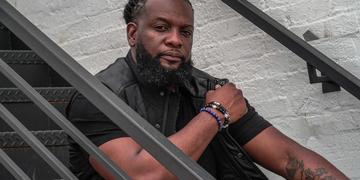 Man with braided hair and beard sitting on stairs wearing a black vest and bracelets.