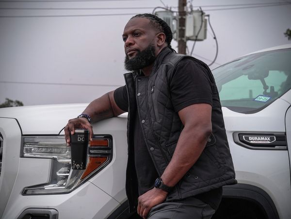 A man leans on a white Duramax truck holding a black tumbler.