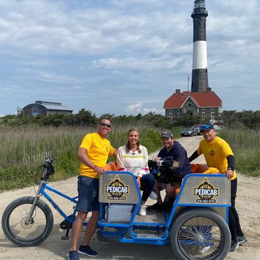 Two men in yellow shirts with a couple on a pedicab near a lighthouse.