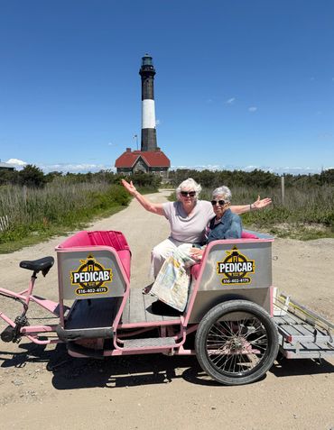 Two women enjoying a pedicab ride near a lighthouse under clear blue skies.
