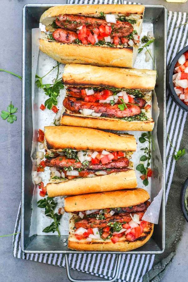 Grilled sausages in baguettes topped with diced tomatoes and onions, served with green herb sauce.
