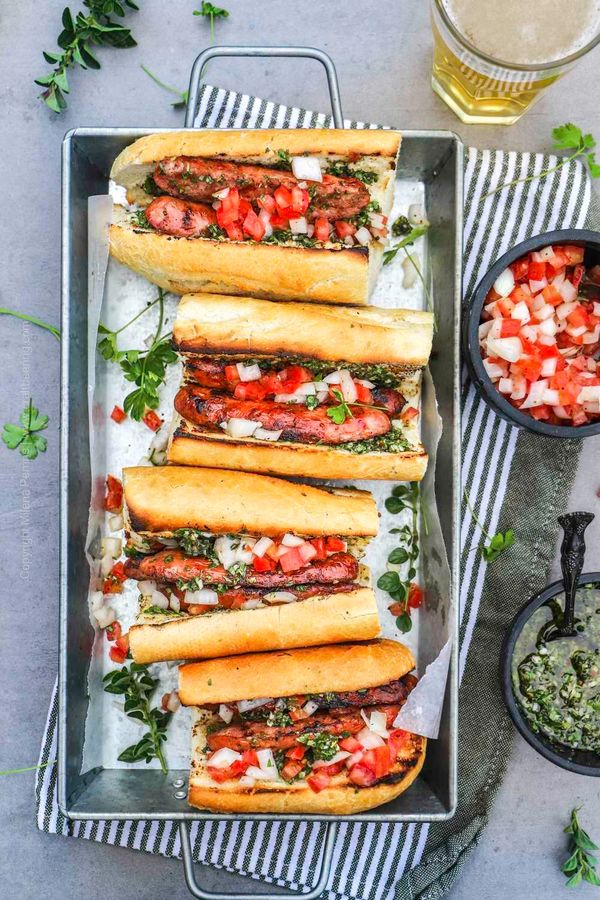 Grilled sausages in baguettes topped with diced tomatoes and onions, served with green herb sauce.