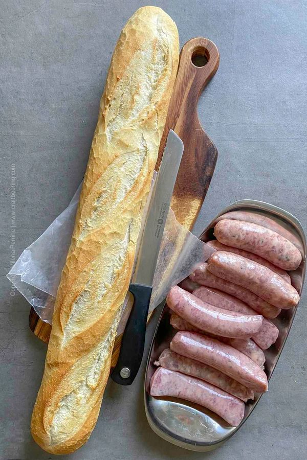 Baguette with a serrated knife and raw sausages on a cutting board.