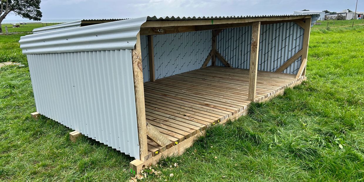 A simple open-sided shelter with a corrugated metal roof and wooden floor in a grassy field.