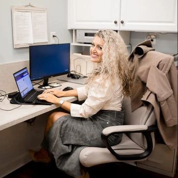 Woman working on a laptop at her office desk, smiling at the camera.