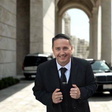 Man in a suit smiling outdoors near cars.