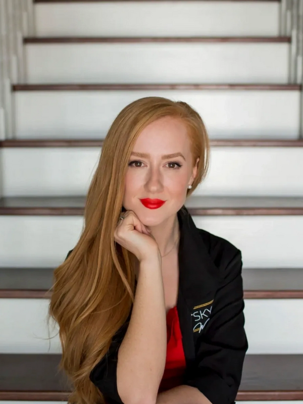 Woman with long red hair sitting on white stairs, wearing a red dress and black jacket.