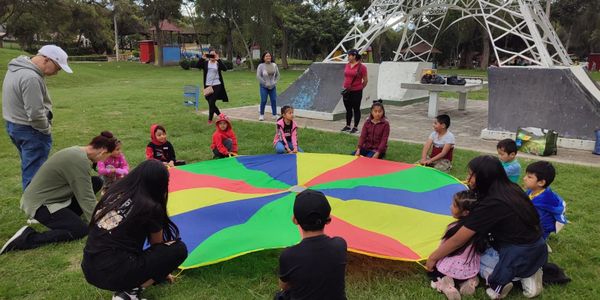 Children and adults gather around a colorful parachute outdoors.