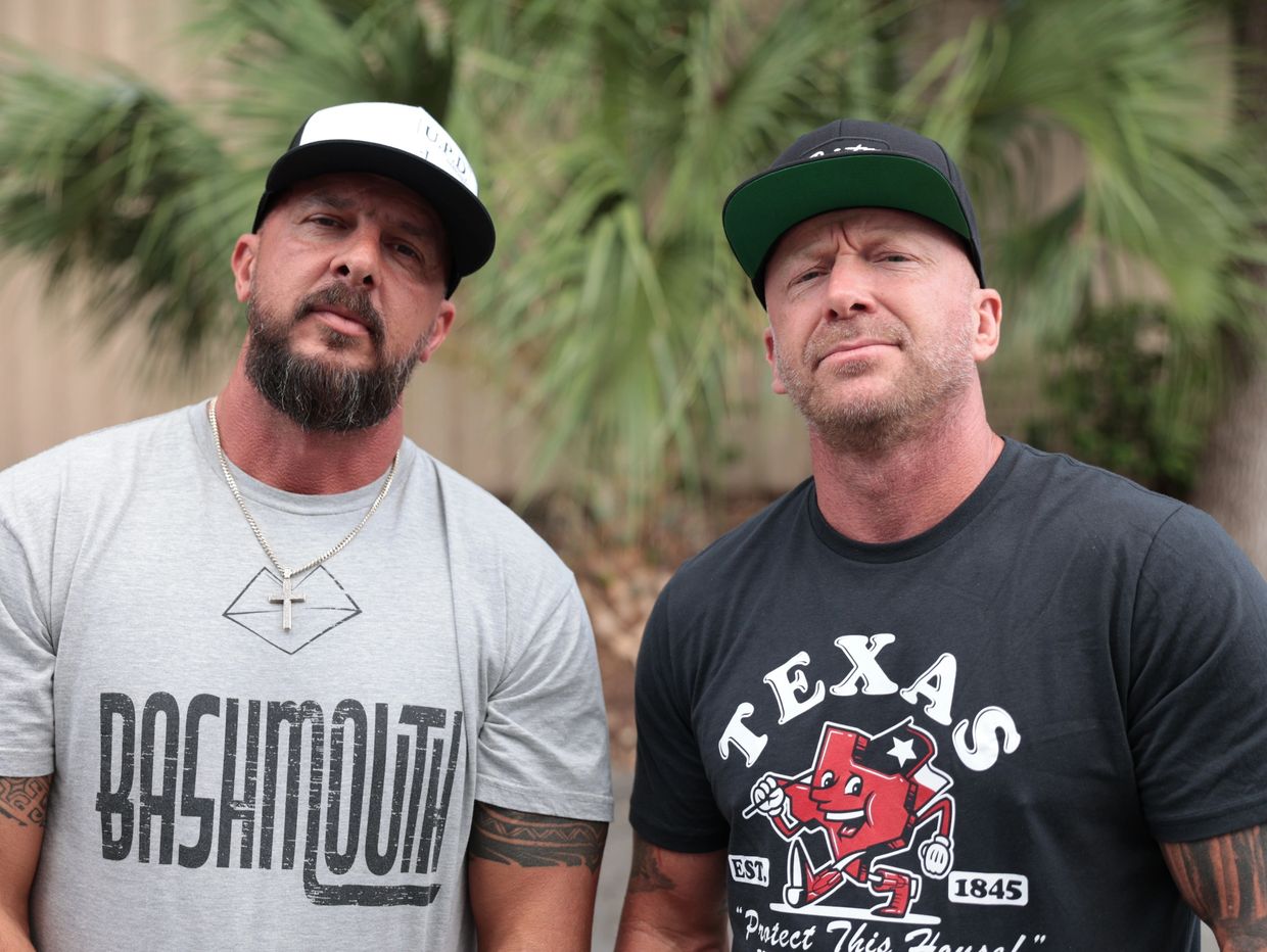 Two muscular men wearing caps and graphic tees pose outdoors with serious expressions.