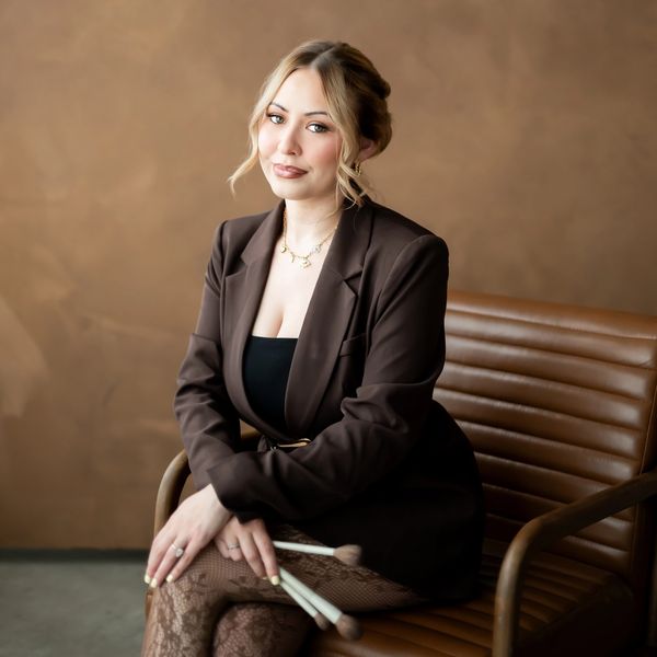 A woman in a brown blazer holding makeup brushes, sitting on a leather chair.