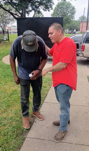 Man in red comforting a distressed man outdoors near a sidewalk.