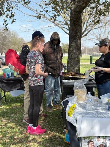 People gathered at an outdoor food stand under a tree on a sunny day.