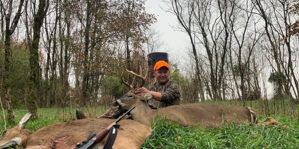 Hunter posing with two deer in a forest clearing.
