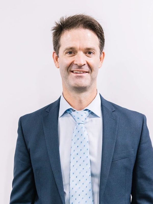 Man in a navy blue suit and light blue patterned tie smiling at the camera.