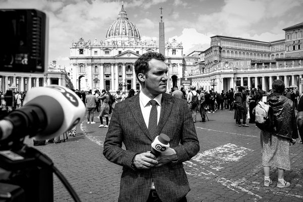 A reporter holding a microphone with a historic building in the background.