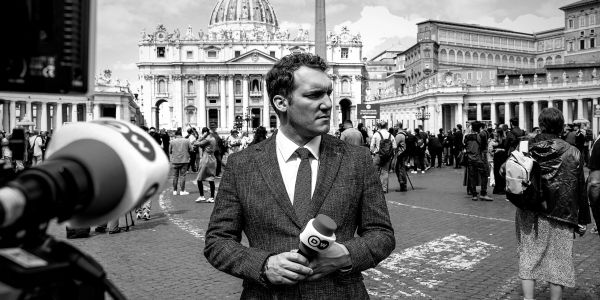A reporter holding a microphone with a historic building in the background.