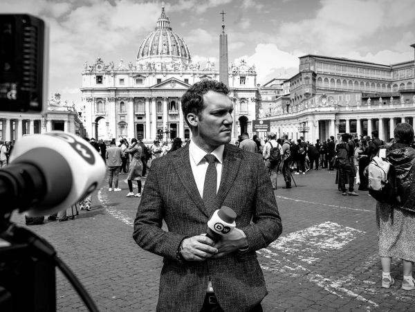 A reporter holding a microphone with a historic building in the background.