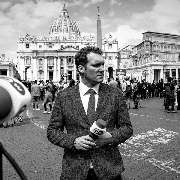 A reporter holding a microphone with a historic building in the background.