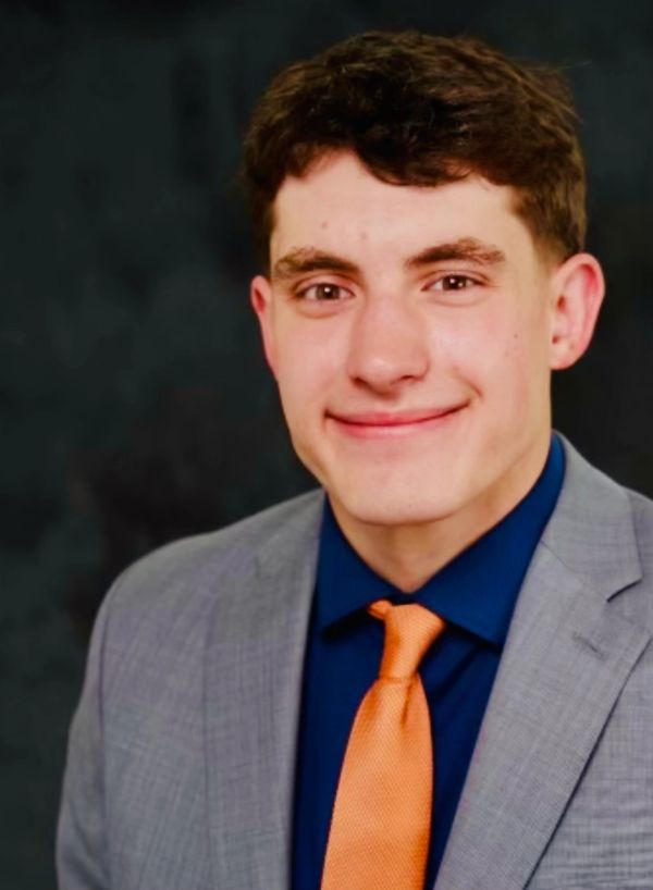 Young man in a gray suit with an orange tie smiling against a dark background.
