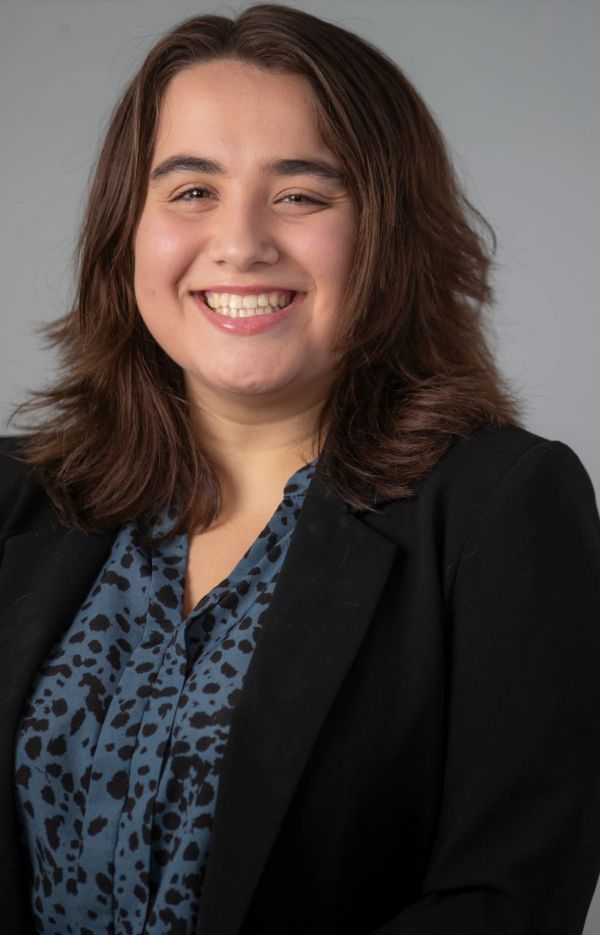 Smiling woman in a black blazer and patterned blue shirt against a gray background.