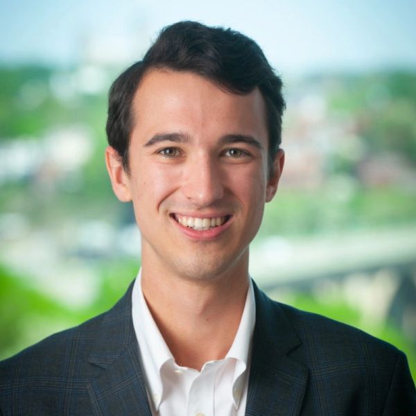 Young man in business attire smiling outdoors with blurred city background.