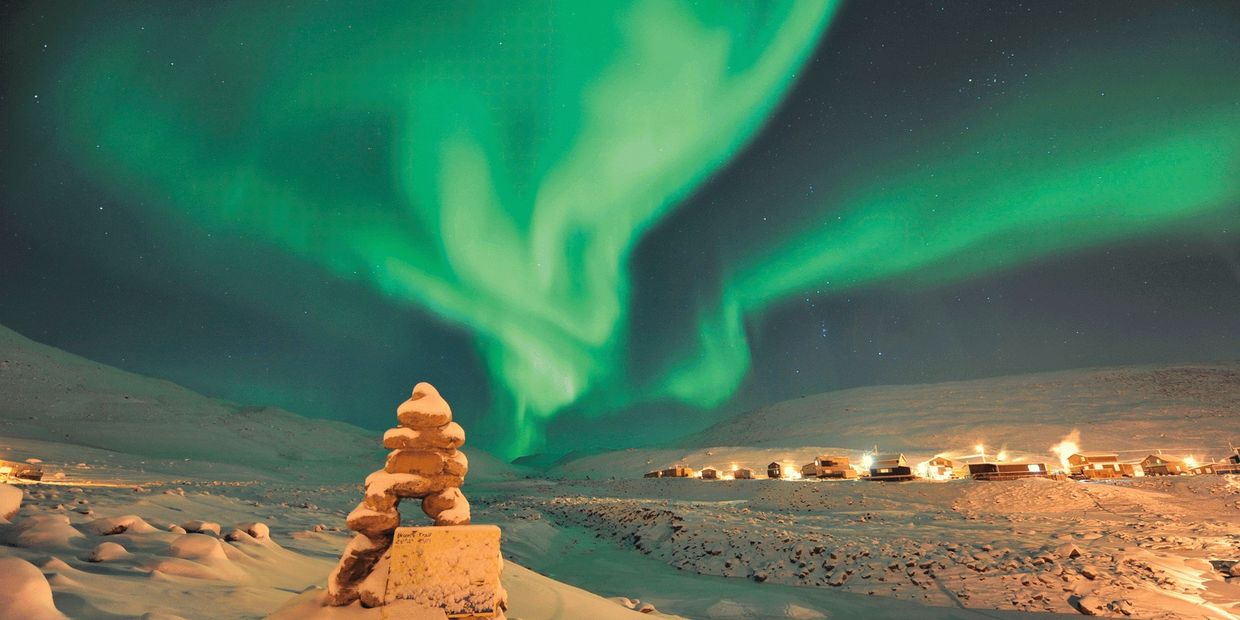 Northern lights glow over snowy landscape and lit village at night.