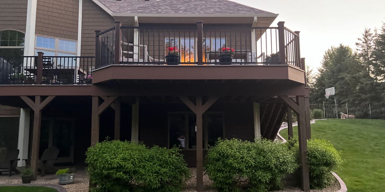 Backyard deck with flower pots and outdoor seating above a garden with bushes.