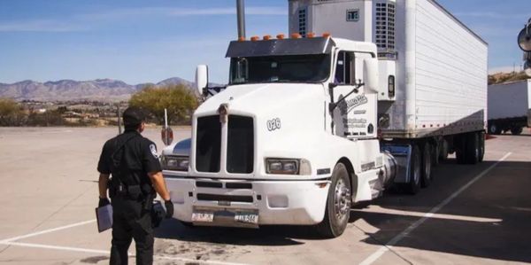 A police officer inspects a white semi-truck parked in a lot on a sunny day.
