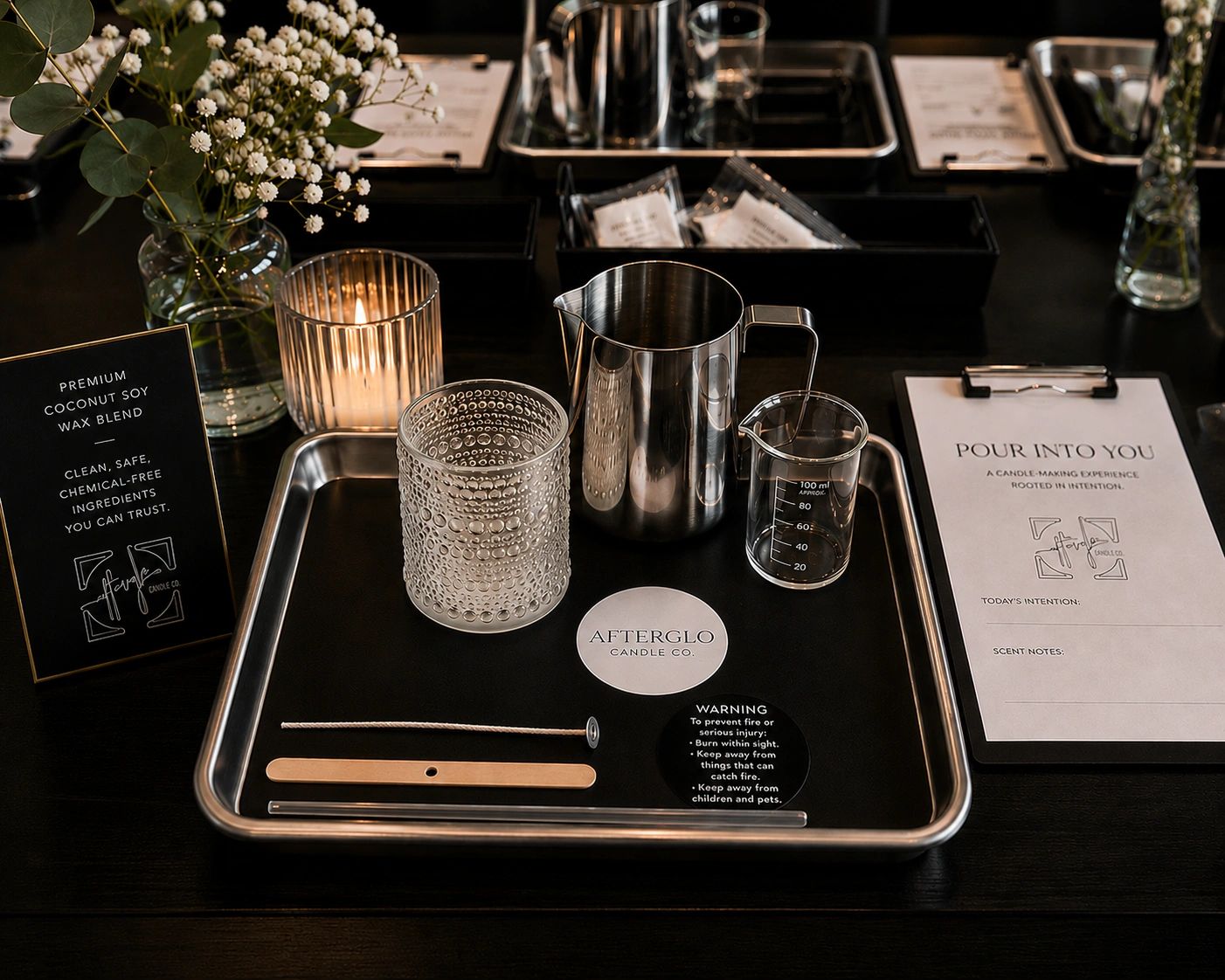 A candle-making kit with glassware, wick, and instructions on a metal tray.