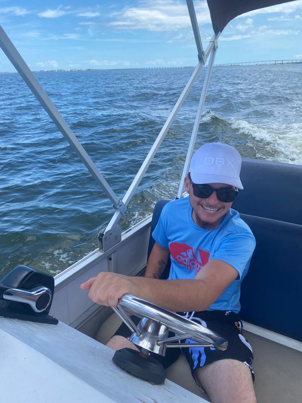 Man steering a boat on a sunny day, smiling with sunglasses and a white cap.