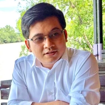 Man in white shirt sitting at a table outdoors with greenery in the background.