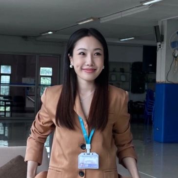 Young woman in a brown blazer smiling indoors with hands in pockets.