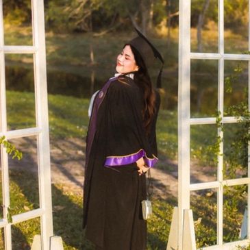 A graduate in black gown and cap posing outdoors near white window frames.