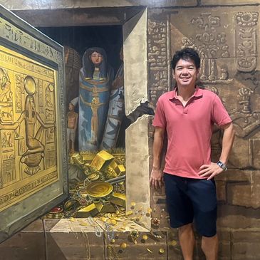Man stands smiling beside a painted Egyptian treasure mural with hieroglyphics and gold.