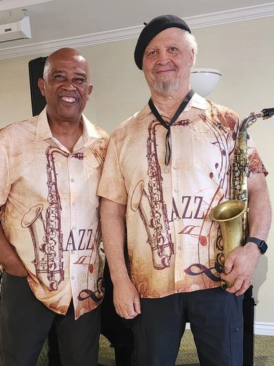 Two men wearing matching jazz-themed shirts, one holding a saxophone.