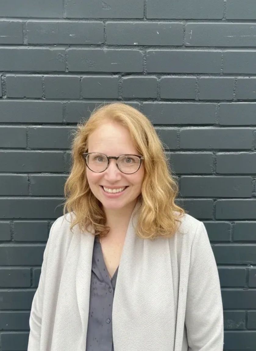 Smiling woman with glasses in front of a gray brick wall.