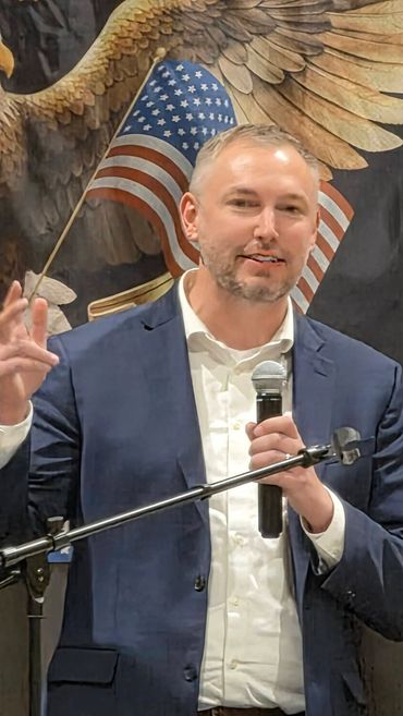 Man in suit holding microphone with American flag and eagle backdrop.
