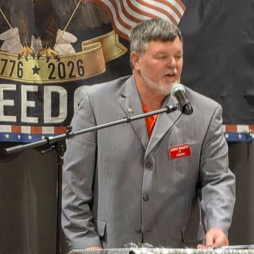 Man in a gray suit speaking at a podium with an American flag backdrop.