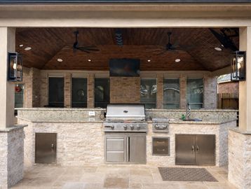 Outdoor kitchen with grill and stone countertops under a wooden ceiling.