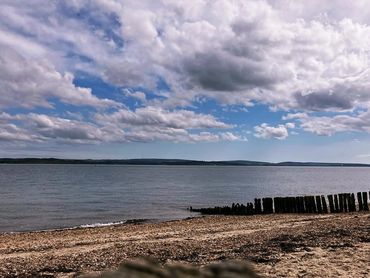 A sandy beach looking out towards a calm sea and land in the distance against a blue cloudy sky.