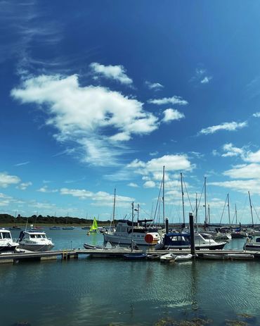 A small marina with a few boats on calm water, backed by a sunny blue sky with a few white clouds.
