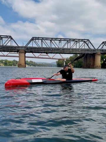Man paddling a red kayak on a river under a steel bridge.