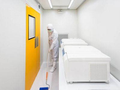 Person in protective suit cleaning a sterile lab with white machines and a bright yellow door.