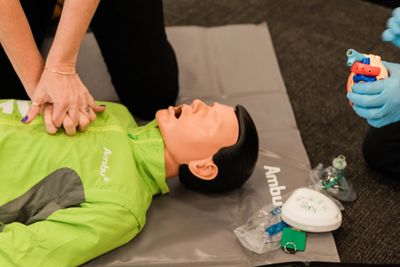 Person practicing CPR on a medical training mannequin with defibrillator nearby.