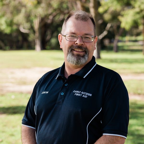 Smiling man wearing a black first aid polo in a sunny park.