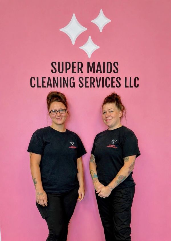 Two women in black uniforms standing against a pink wall with cleaning service logo.