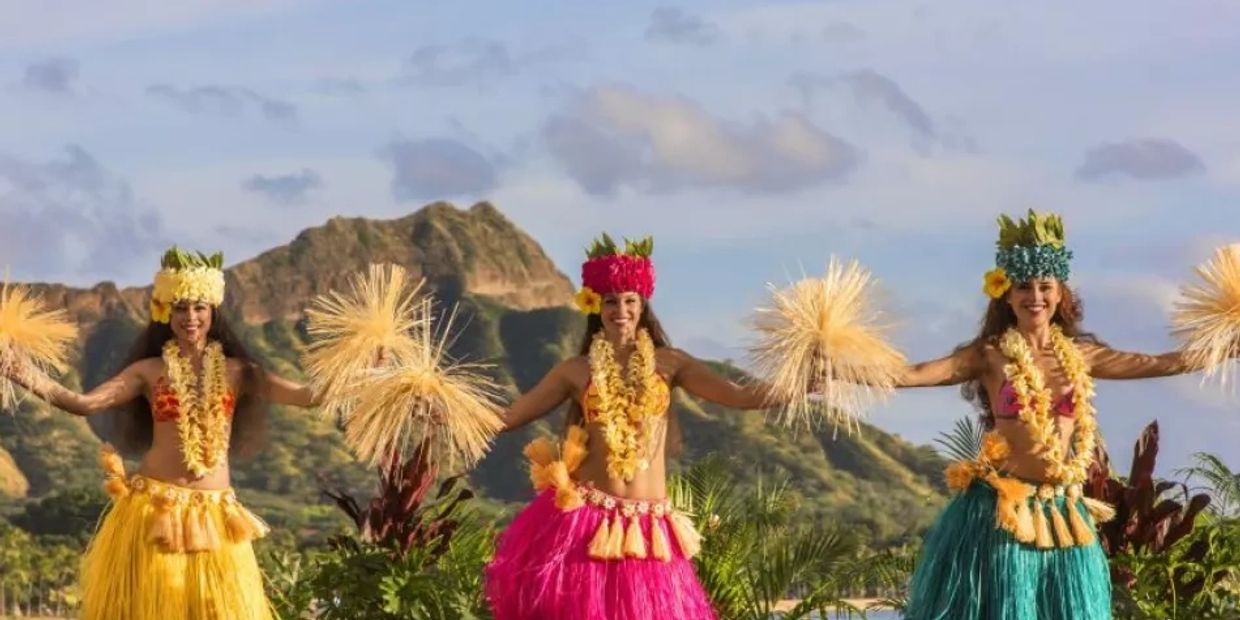 Three hula dancers performing outdoors with mountain backdrop.