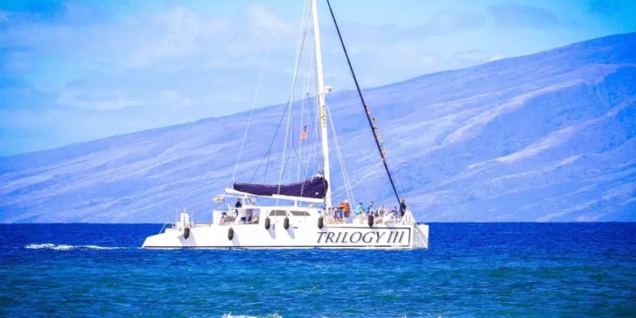 Sailboat named TRILOGY III on blue ocean with mountain backdrop.