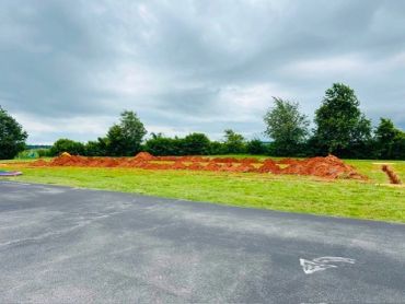 A long trench dug in a grassy area near a paved road under a cloudy sky.
