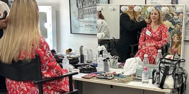 Woman in red dress getting makeup done in front of a mirror.